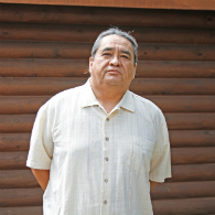 Photo of Manley Begay Jr. standing in front of a wood log wall.