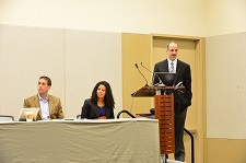 Photo of a male standing at a podium with a female and another male seated next to him at a table.