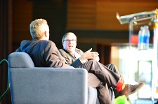 Two males speaking while sitting in sofa chairs which appear to be on a stage.