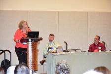 Female wearing a pink top speaking from a podium while two males are sitting at a table to her side.