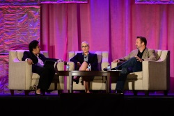 Three people sitting in cushioned chairs on a stage with a pinkish-purple stage curtain behind them.