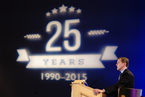 Male speaking from behind a podium with a projection celebrating 25 years visible in the background.