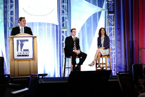 One person standing behind a podium on a stage with two other people sitting in high chairs behind them.