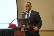 Photo of a male of color dressed in a suit while speaking from behind a podium.