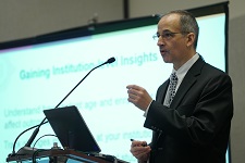 Male presenter speaks from a podium with slides being projected on a screen behind him.