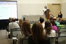 Audience members watch someone in a white shirt speak from a podium at the front of the room.