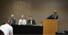 Panel of three people sitting at a table as one more figure stands at a podium next to them.