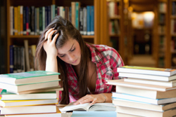 Female student appearing to be stressed as she reads a book with others piled next to her.