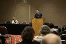 Male in a dark colored polo shirt speaking from behind a podium.