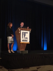 Photo of two people, a male and female, standing behind a podium on a stage.