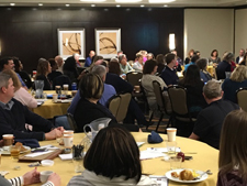 Event space full of people sitting at circular tables with plates of food visible. 