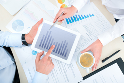 Two sets of hands collaborate over a tablet with a bar chart visible on its display.