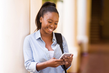 Female of color smiling at her phone while wearing a backpack over one shoulder.