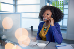 Female of color speaking on a phone call while sitting at an office desk with a large computer monitor.