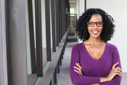 Female of color wearing a purple top smiling while standing with her arms folded.