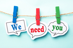 The phrase "we need you" written on pieces of paper hung by colorful clips on a string.