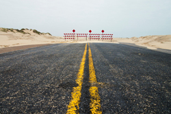 Photo of an asphalt road through a desert with the double-yellow line extending into the distance where a road block is visible.