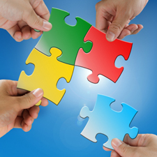 Four hands, each with its own puzzle piece, work together to solve the puzzle.