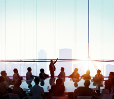 Photo of a board meeting taking place in a large open room with windows showing a view of skyscrapers. 