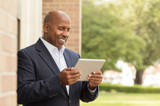 Male of color smiling at a tablet while leaning against the outside of a building.