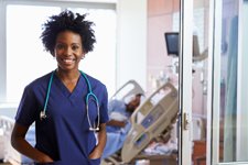 Nurse of color smiling in front of the door to a patient's room.