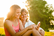 Mother and daughter smiling at a phone together as they sit outside in the grass on a sunny day.