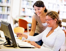 Two females smiling as they work on a computer together in what appears to be a library.