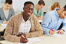 Student of color smiling at the camera while they and the other students take an exam. 